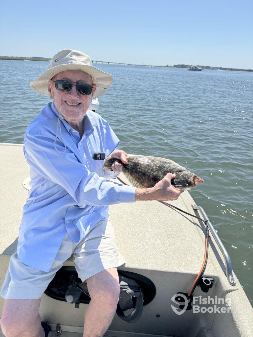An elderly angler proudly displaying a Flounder while fishing on a sunny day, showcasing the joy of fishing in a coastal environment.