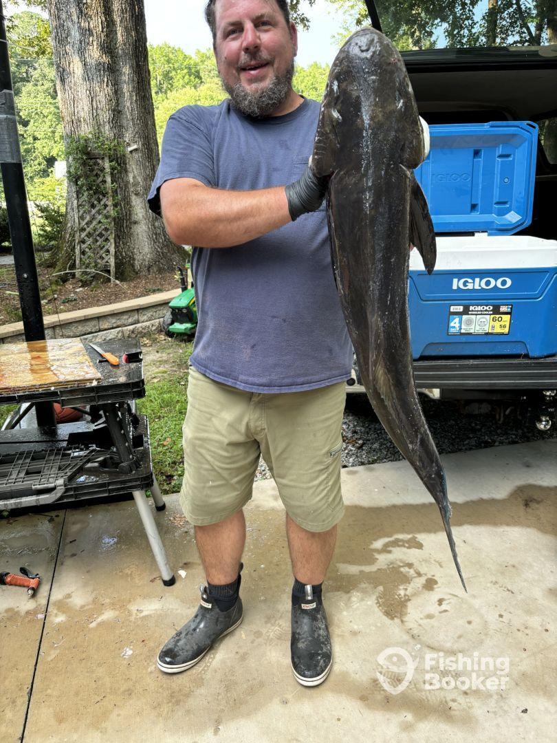 An angler proudly displaying a large Catfish after a successful fishing trip, with a cleaning station visible in the background.