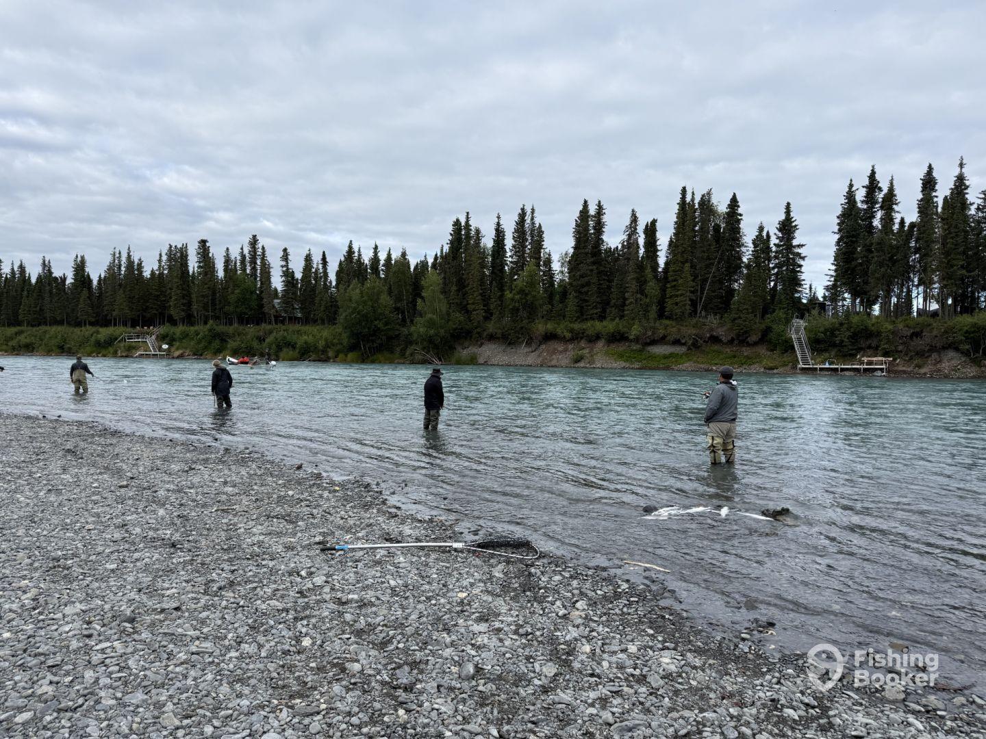 Group of anglers wading in a river, casting lines in a scenic Alaskan fishing environment surrounded by lush trees.