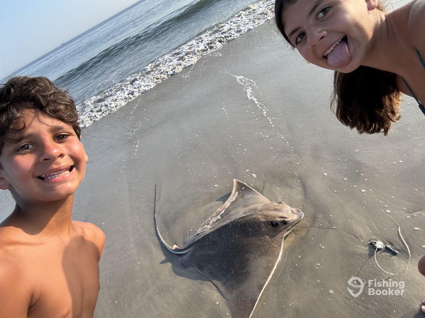 Two young anglers posing with a caught stingray on the beach, showcasing their excitement after a fun fishing adventure.