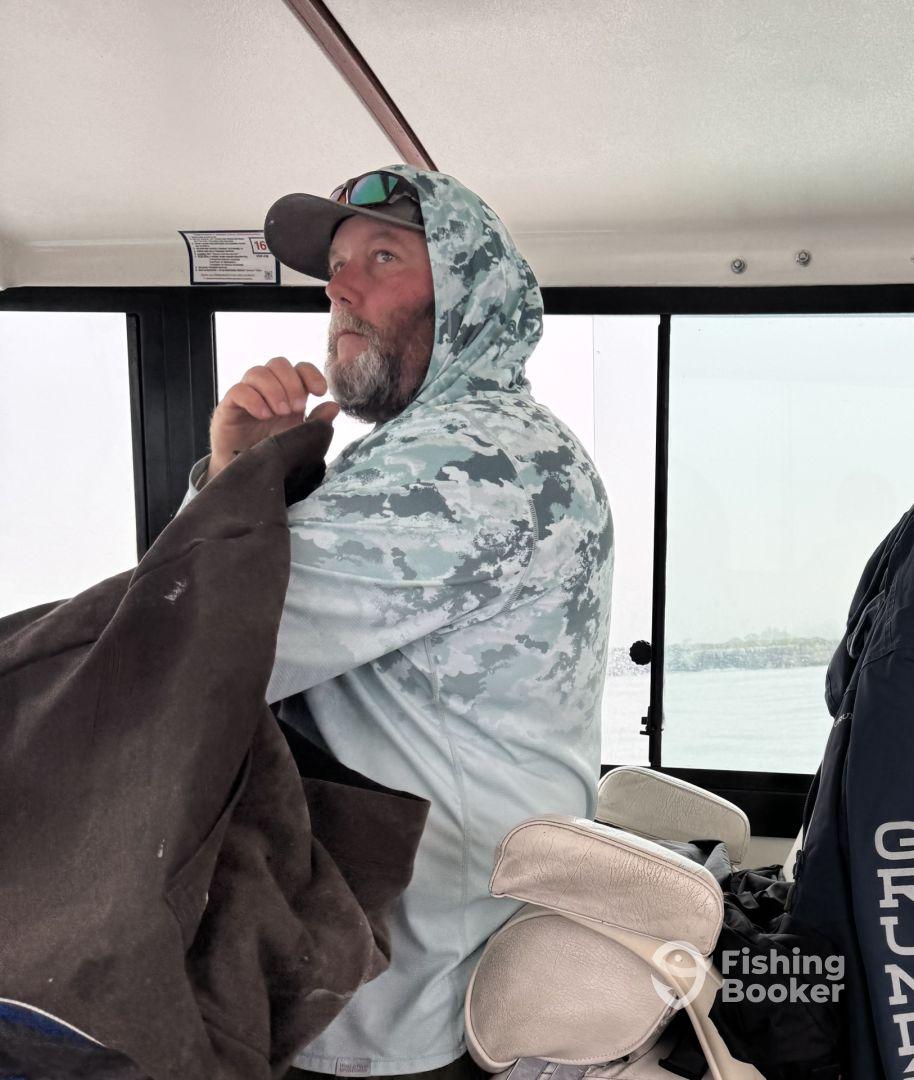 A fisherman inside the cabin of a boat, wearing a camo hoodie and looking thoughtfully out the window, suggesting a moment of anticipation during a fishing trip.