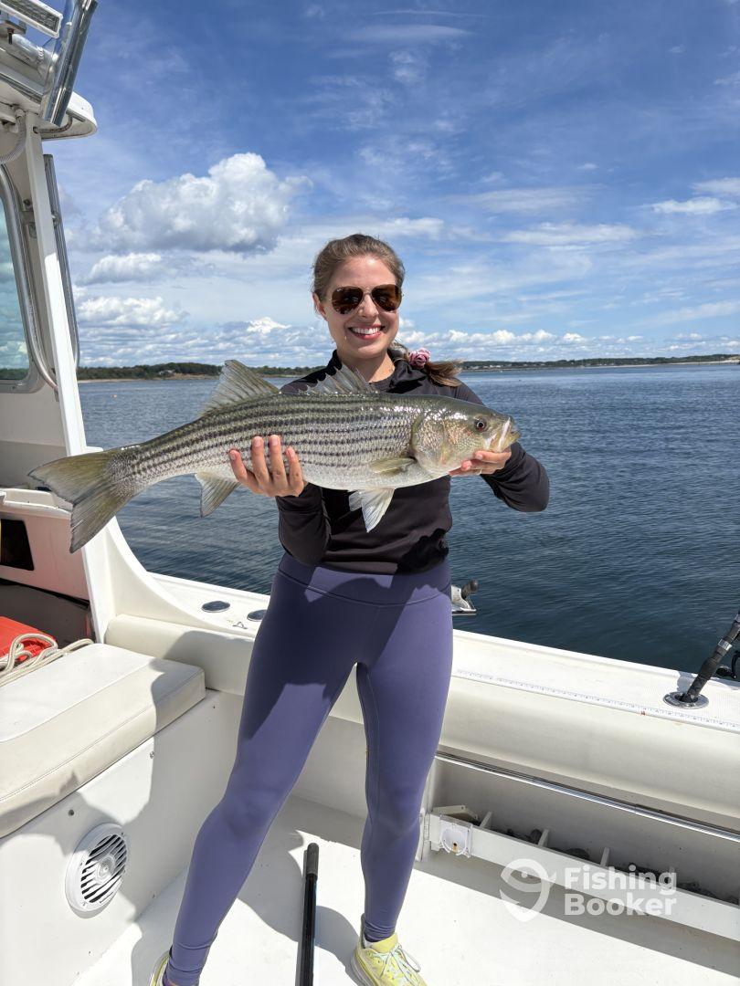 An angler proudly displaying a large Striped Bass aboard a fishing boat, enjoying a sunny day on the water.