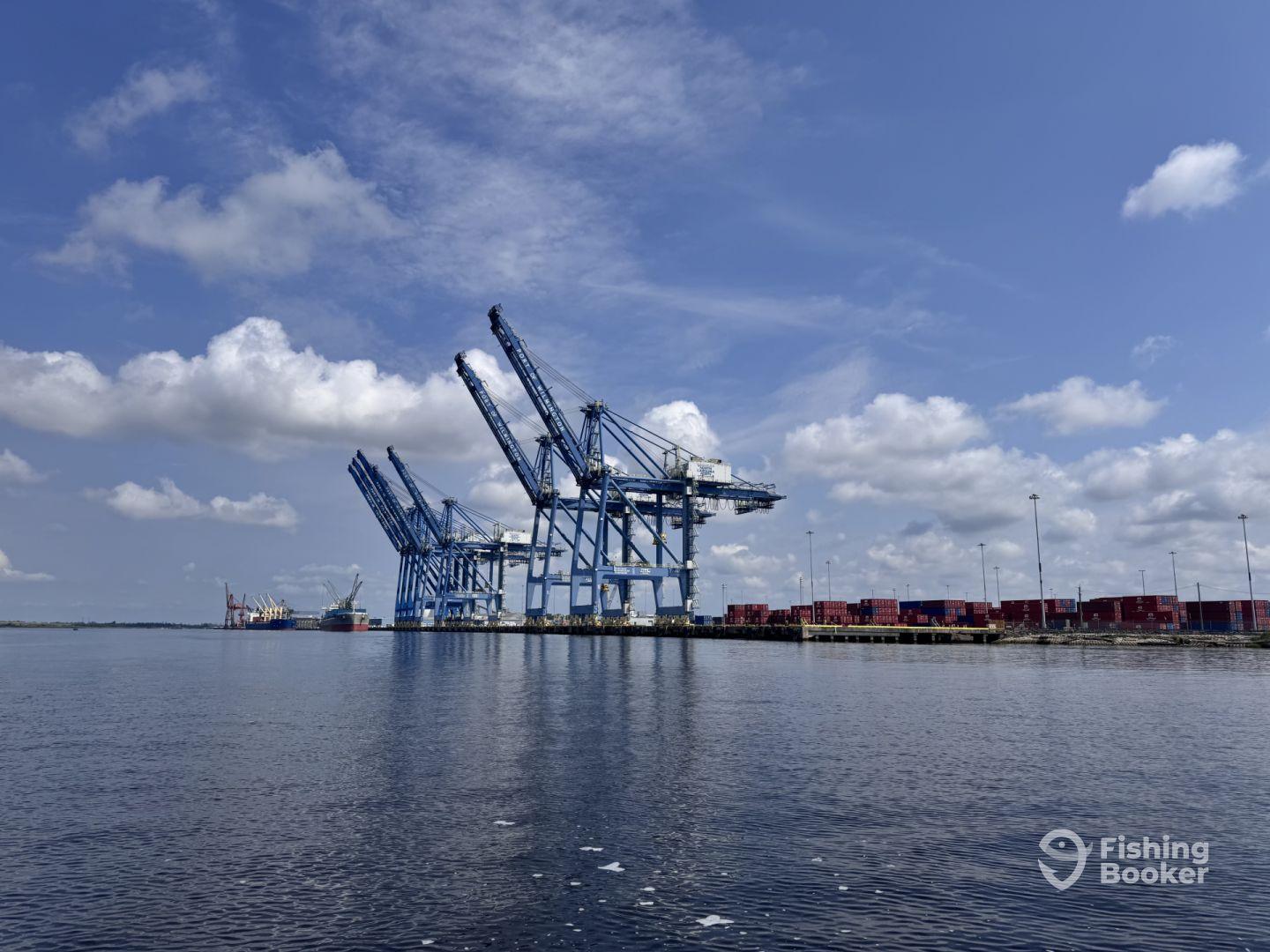 A view of industrial cranes and shipping containers at a port, with a clear blue sky and calm water.