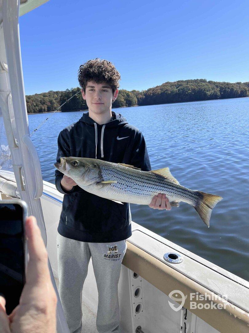 A young angler proudly displaying a large Striped Bass while fishing on a serene lake, surrounded by autumn foliage.
