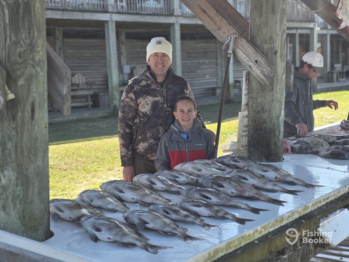 A proud father and son stand beside a neatly arranged display of freshly caught fish at a fishing dock, showcasing their successful day on the water.