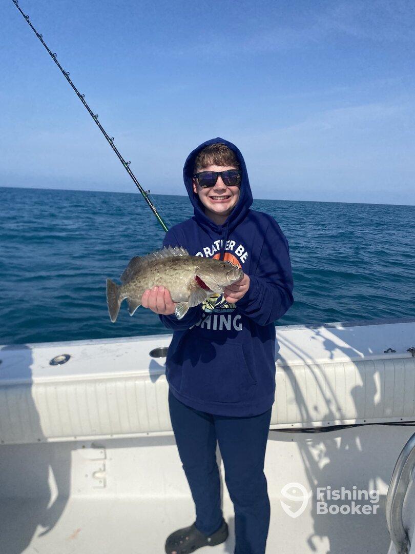 A young angler proudly displaying a large Grouper while fishing on a sunny day at sea.