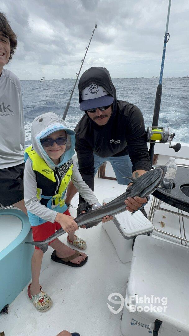 A young angler, wearing a life jacket, proudly holds a fish with the help of an adult on a fishing boat, showcasing a fun family fishing experience.
