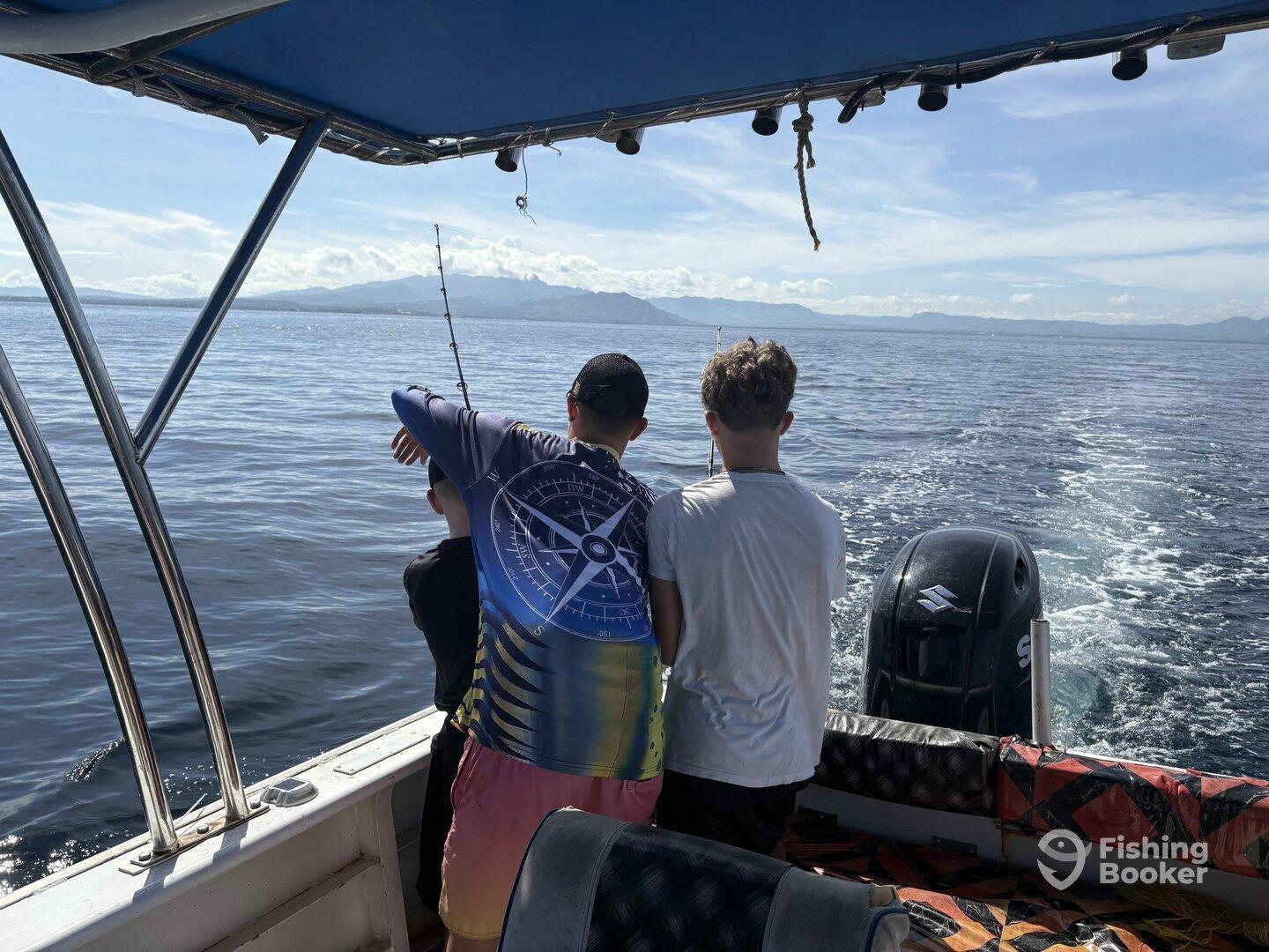 Two young anglers are actively fishing from a boat, enjoying a day on the water with a scenic backdrop of mountains and clear skies.