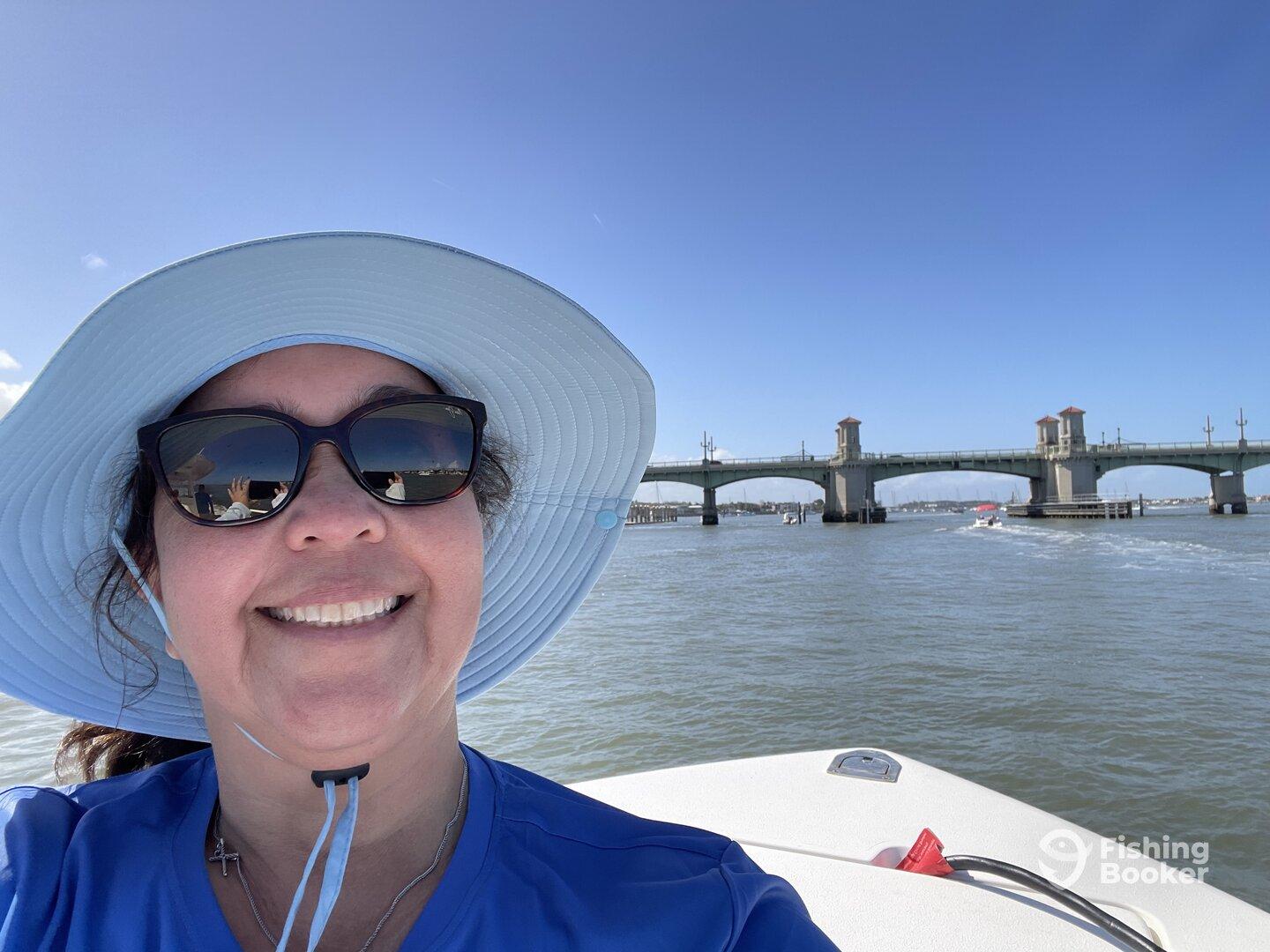 A smiling woman wearing sunglasses and a wide-brimmed hat takes a selfie on a boat, with a bridge visible in the background.