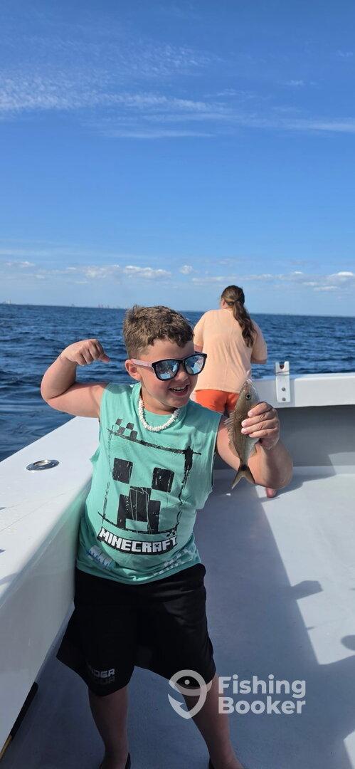 A young angler proudly displaying a small fish while fishing on a boat, with a scenic ocean backdrop.