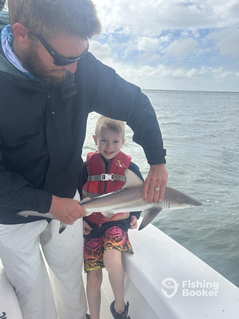 A young boy in a life jacket smiles while holding a small shark, assisted by an adult, on a fishing boat.
