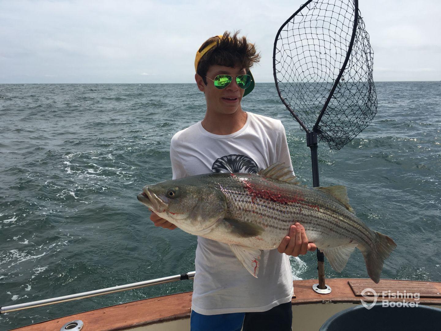 A young angler proudly displaying a large Striped Bass while fishing on a boat in open waters.