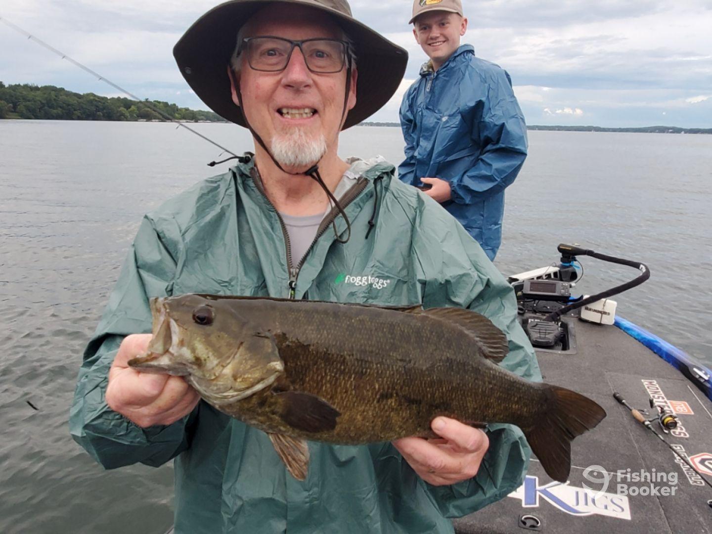 An angler proudly displaying a large Smallmouth Bass while fishing on a calm lake, accompanied by a young companion.