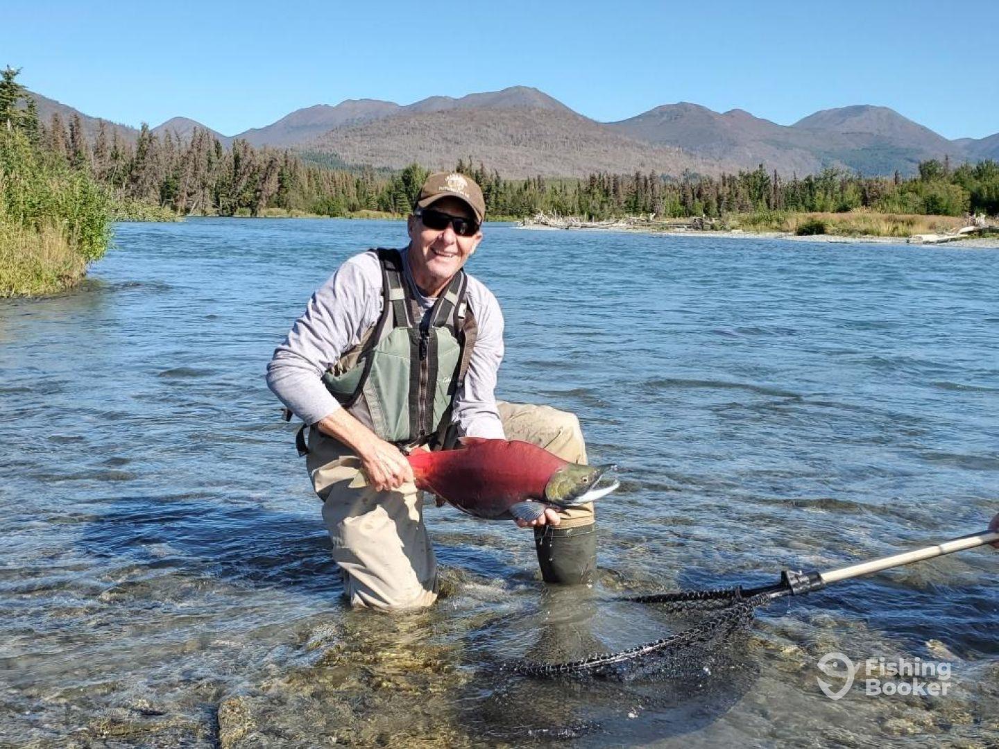 An angler proudly displaying a vibrant Sockeye Salmon while wading in a clear river surrounded by mountainous terrain.