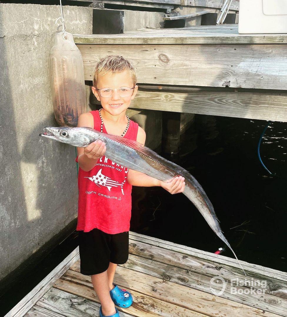 A young angler proudly displaying a long fish, likely a barracuda, at a dock, showcasing a successful fishing trip.