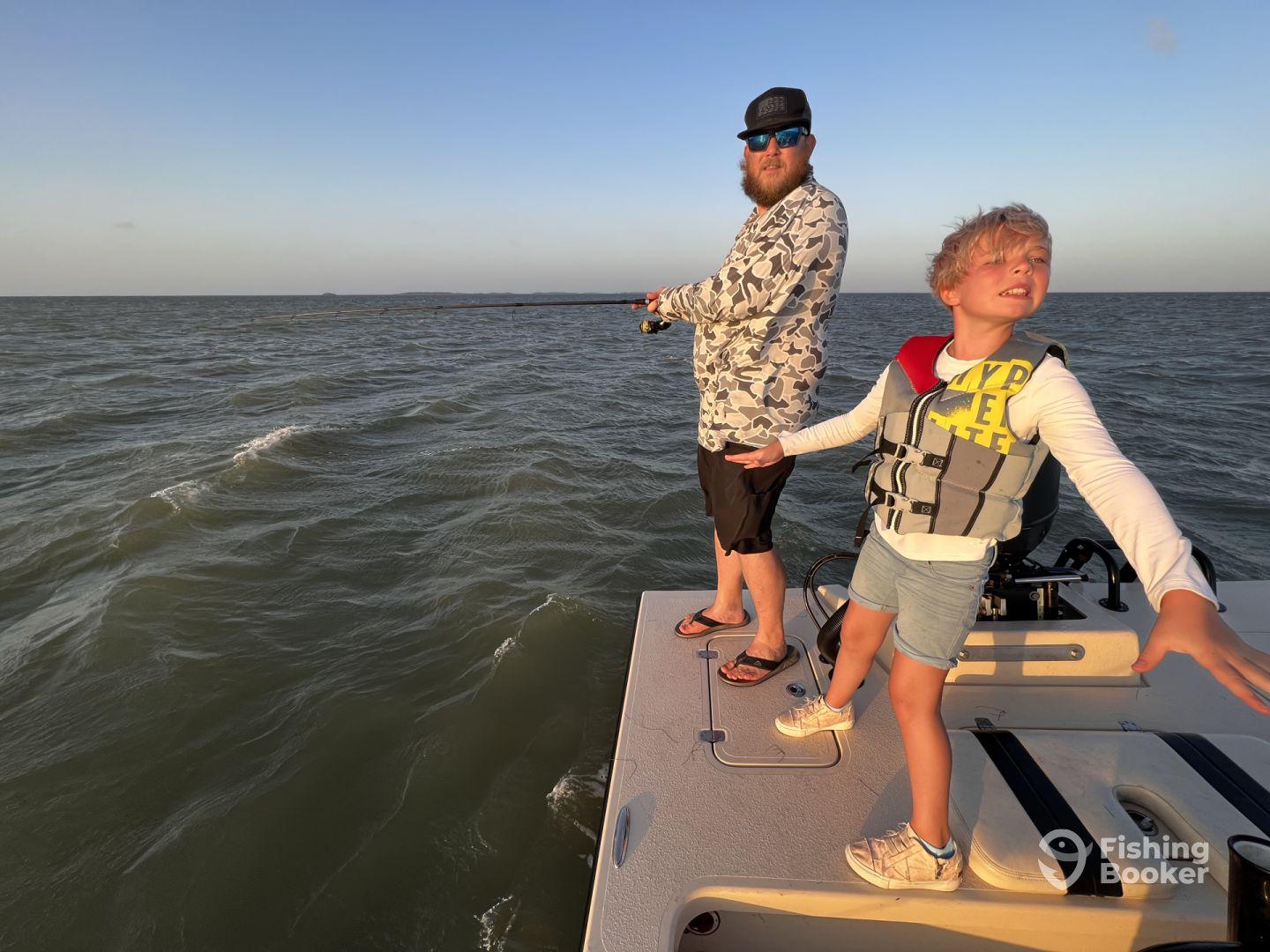 A young angler and an adult are fishing together on a boat, with the child playfully posing while the adult focuses on casting in a scenic fishing environment.