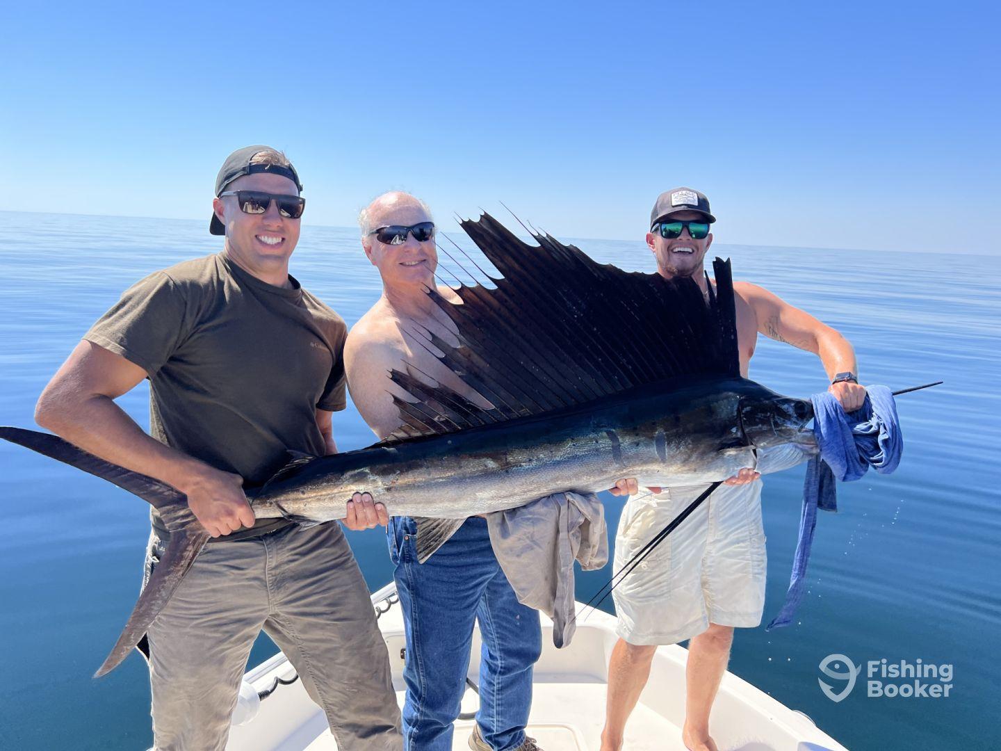 Three anglers proudly displaying a large Sailfish on a calm day at sea, showcasing their successful catch during a deep-sea fishing trip.