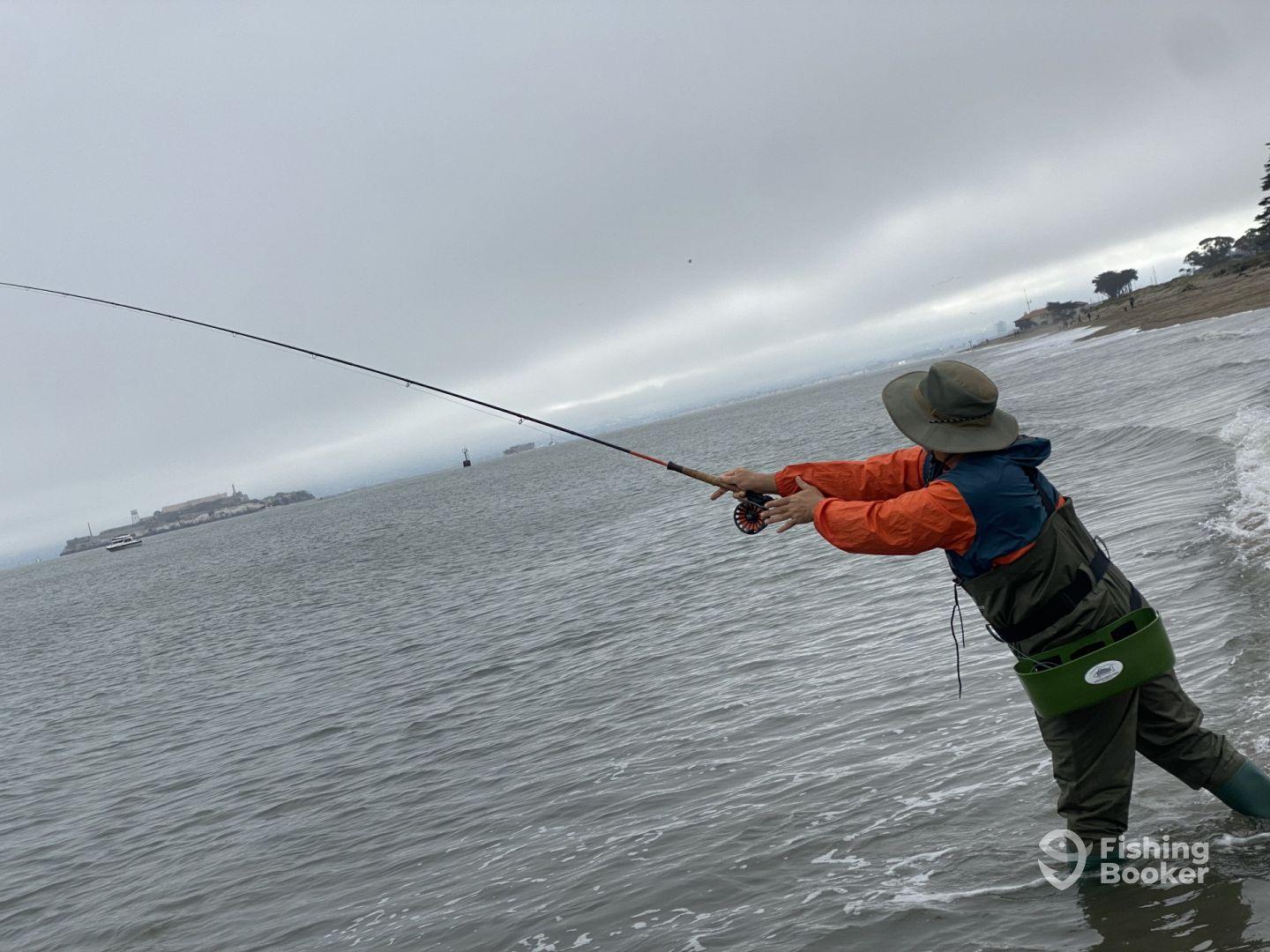 An angler casting a line from the shore with Alcatraz Island in the background, showcasing a cloudy day fishing experience.