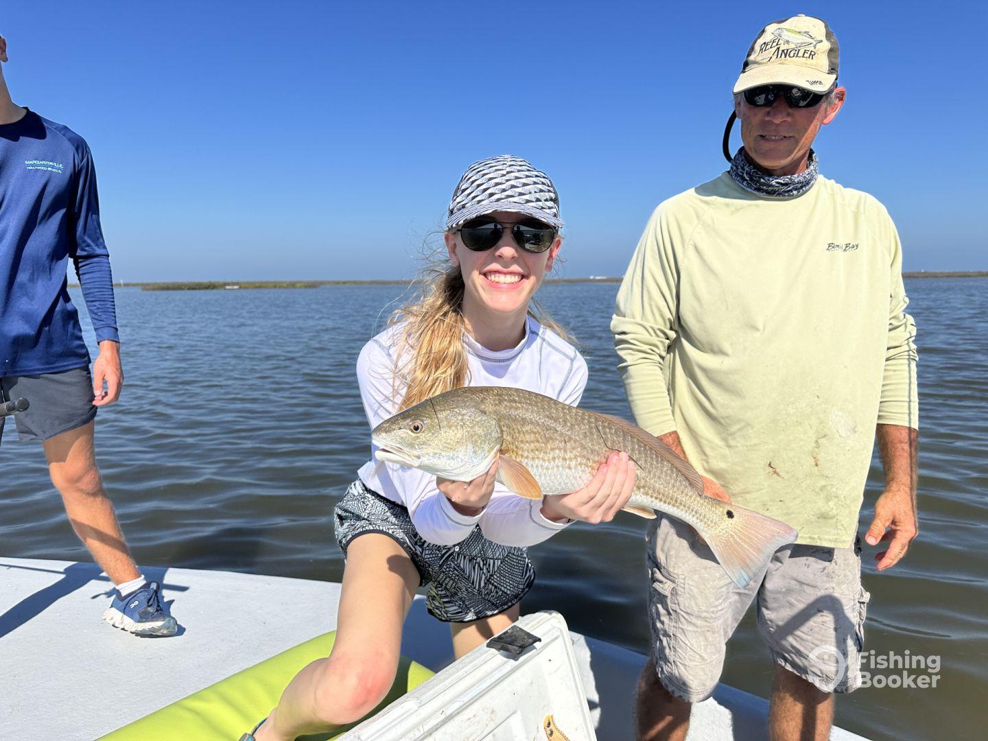 A young angler proudly displaying a Redfish while fishing on a sunny day, surrounded by fellow anglers on a boat.