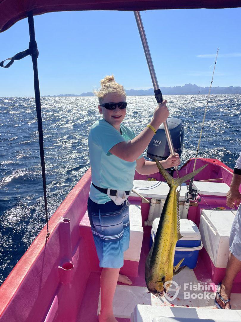 A young angler proudly holding a vibrant Mahi Mahi aboard a colorful fishing boat in sunny waters.
