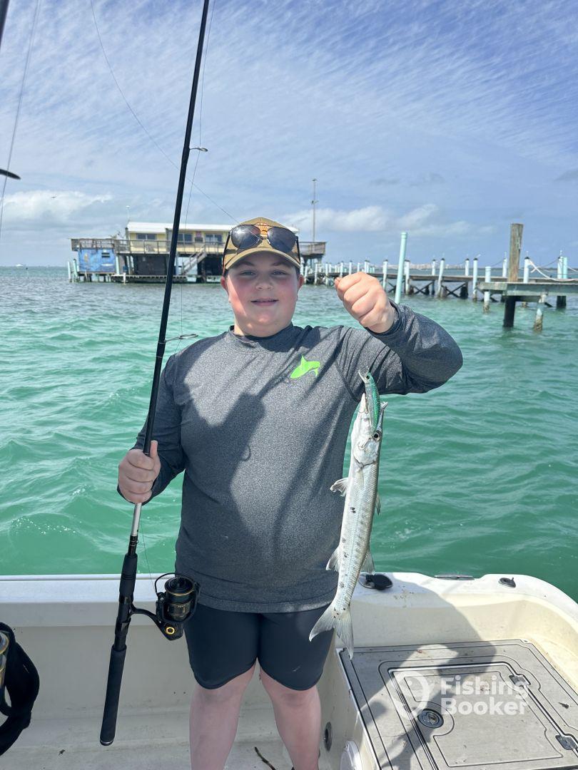 A young angler proudly displaying a Barracuda while fishing from a boat in clear waters.