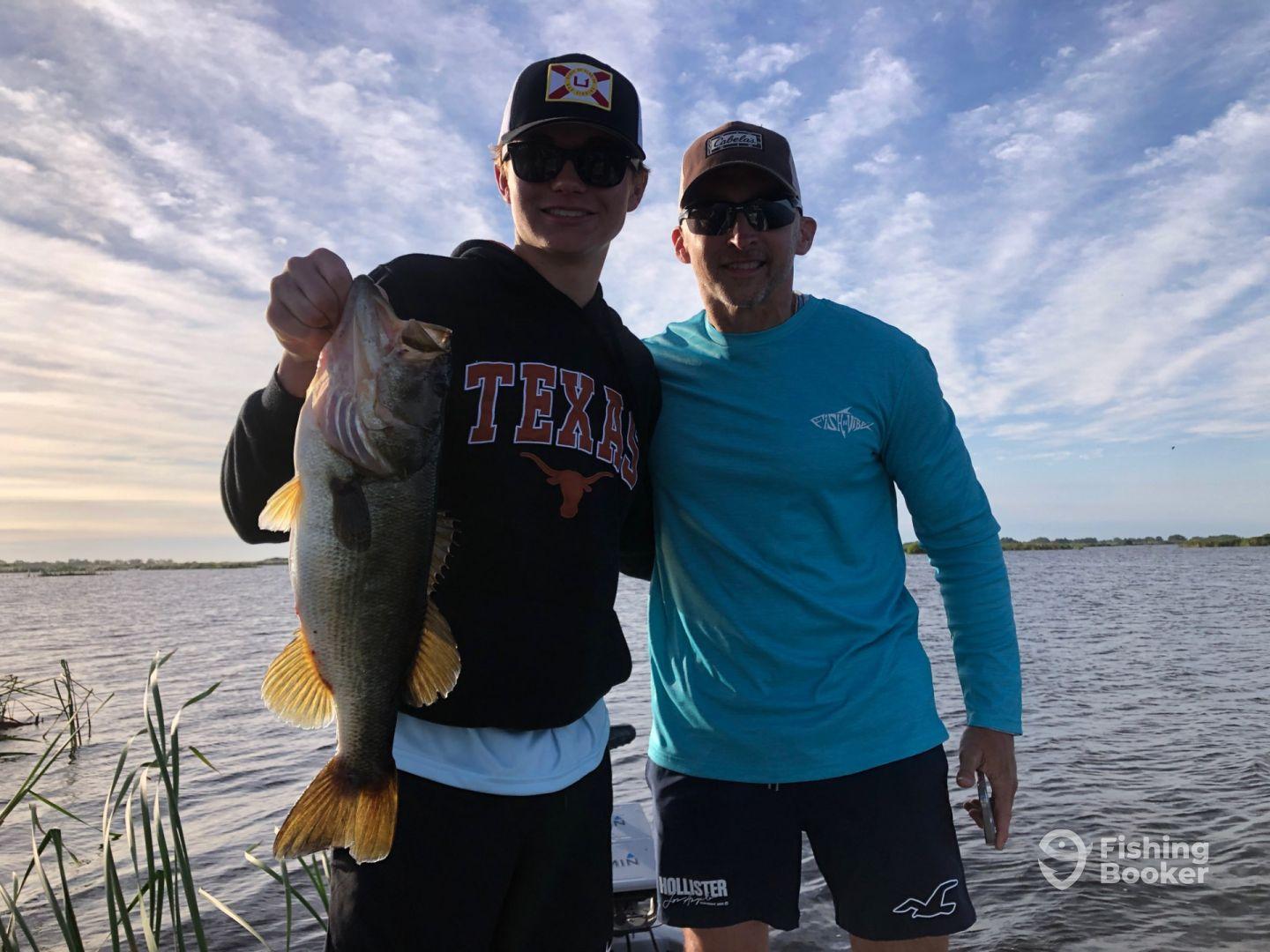 A young angler proudly displaying a large Largemouth Bass while fishing on a serene lake, accompanied by an adult.