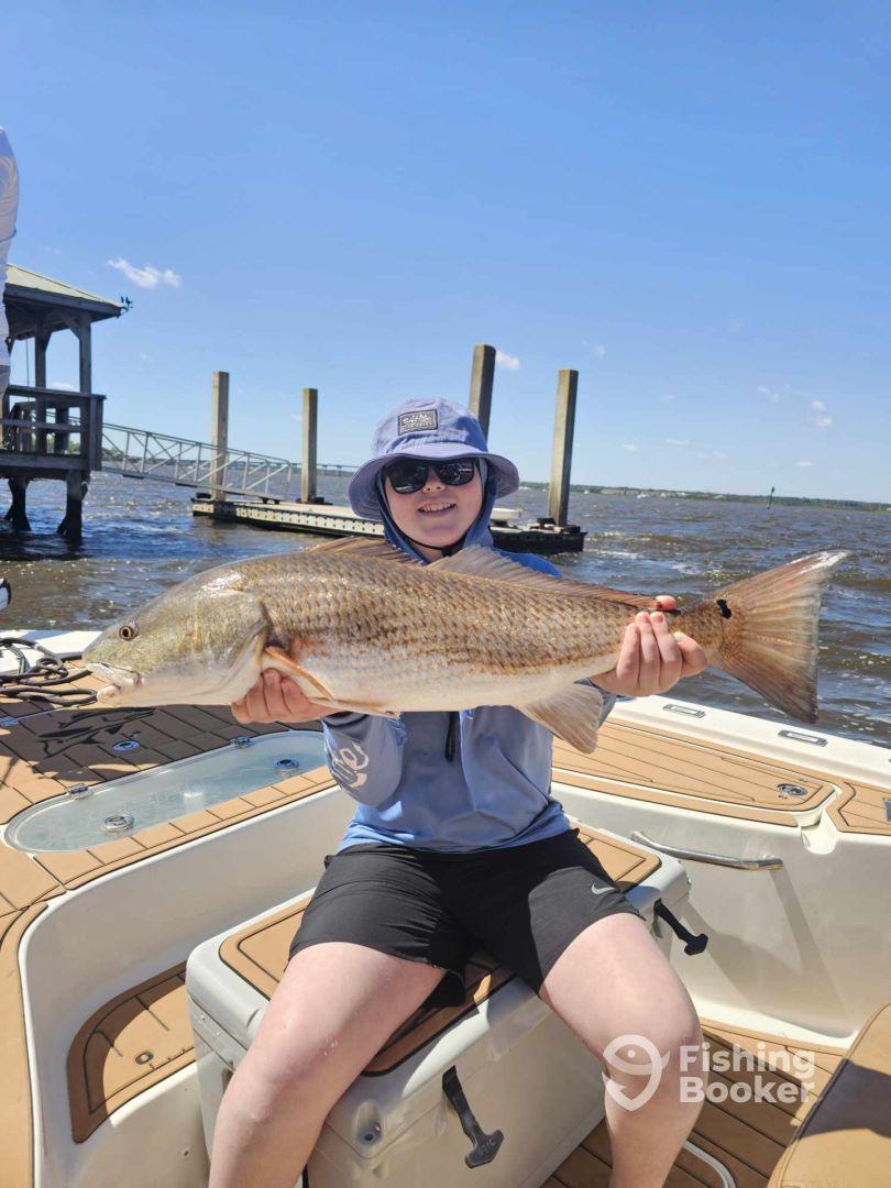 A young angler proudly displaying a large Redfish while seated on a boat, enjoying a sunny day on the water.