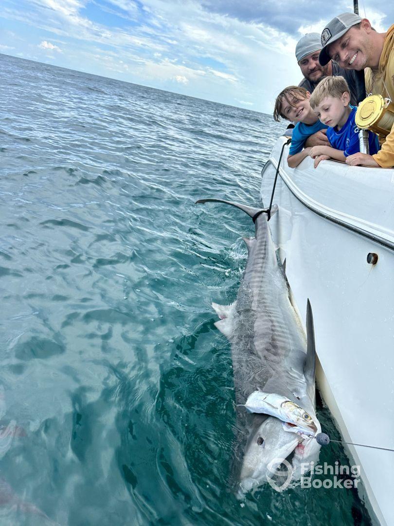 A family of four enjoying a thrilling moment while reeling in a large shark during their fishing trip in open waters.