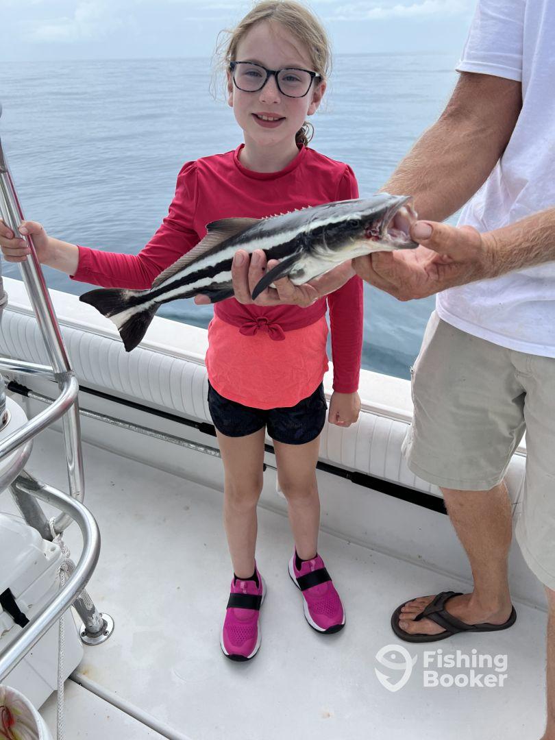 A young girl proudly displaying her catch, a striped fish, while fishing on a boat in calm waters.