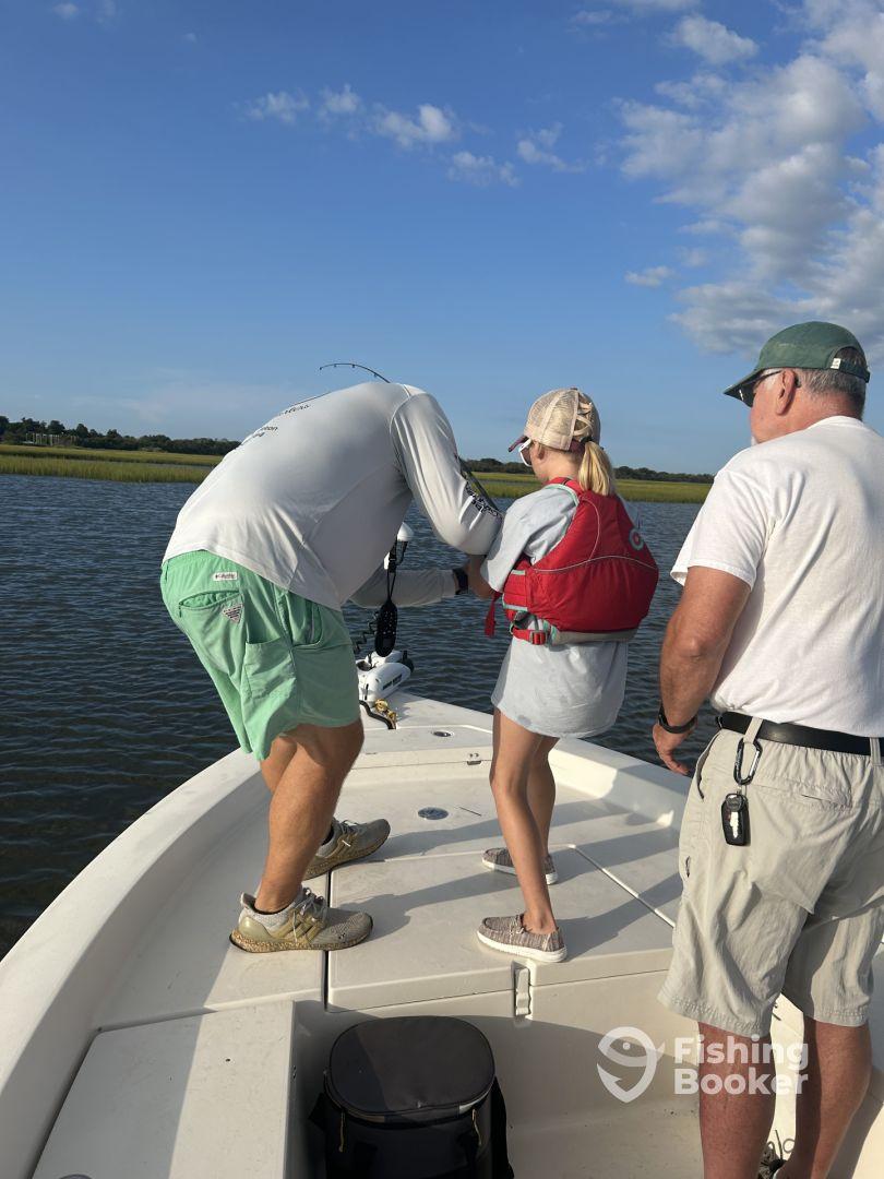 A young girl in a life jacket is being assisted by an adult while reeling in a fish on a sunny day, showcasing a family fishing experience.