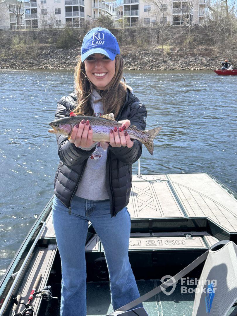 An angler proudly displaying a Rainbow Trout while fishing from a boat on a scenic river.