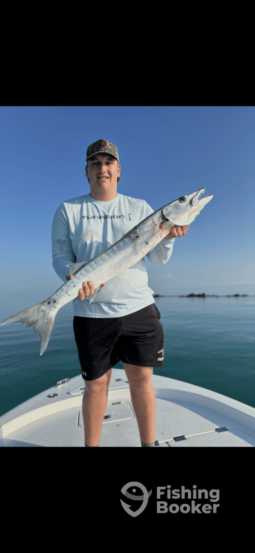 Angler proudly displaying a large Barracuda while standing on the bow of a boat, with calm waters in the background.