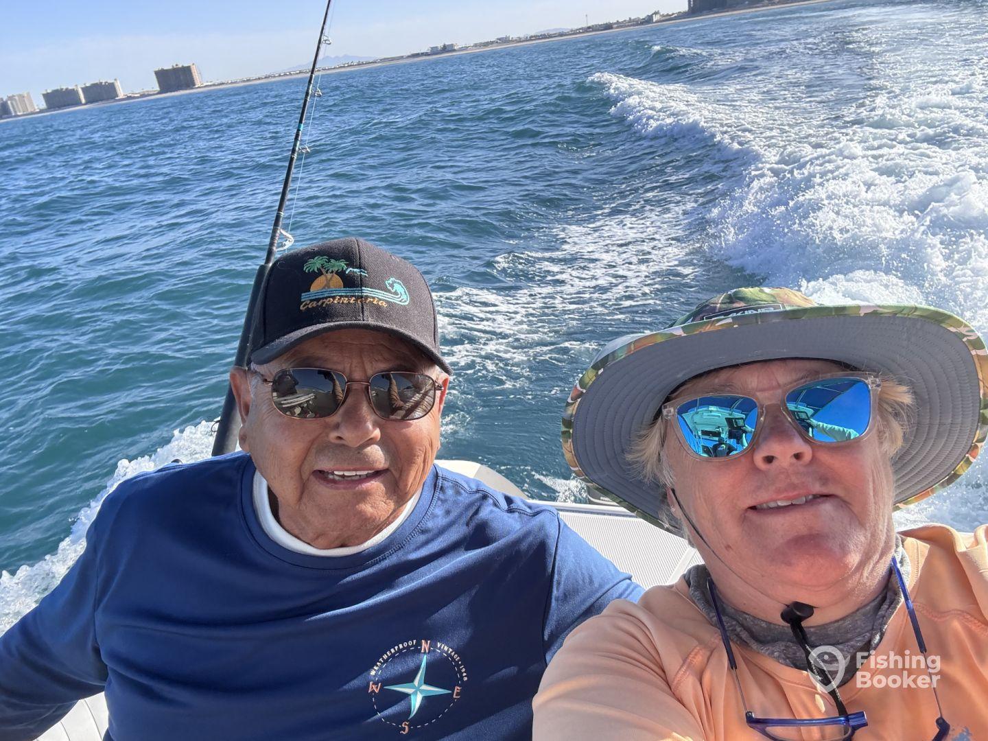 Two anglers enjoying a day on the water, seated inside the boat with a scenic coastal backdrop.