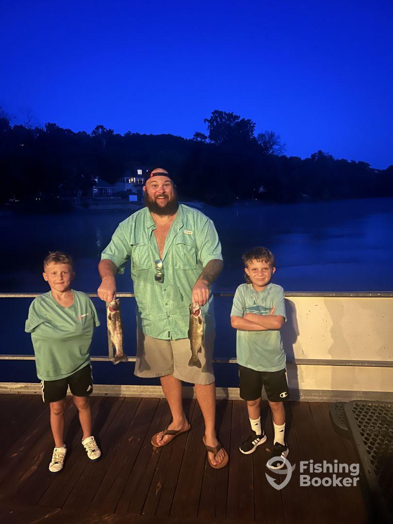 A proud adult and two young anglers displaying their catch of freshwater fish at dusk by the water.
