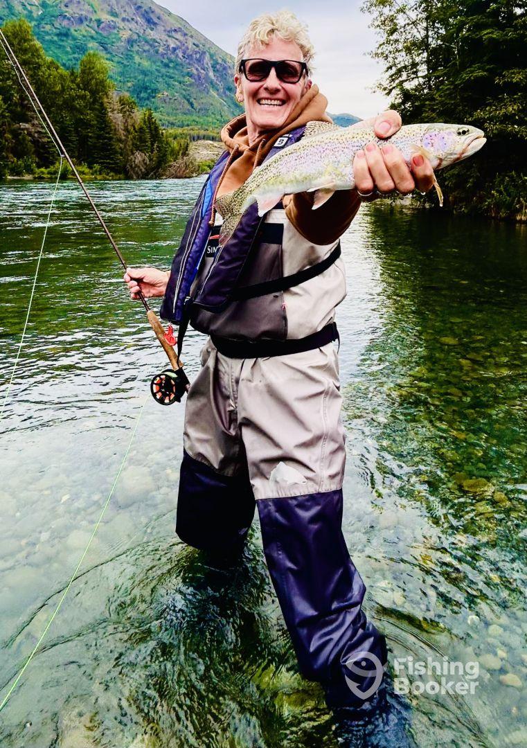 An angler proudly displaying a vibrant Rainbow Trout while wading in a clear river, surrounded by lush greenery and mountains.