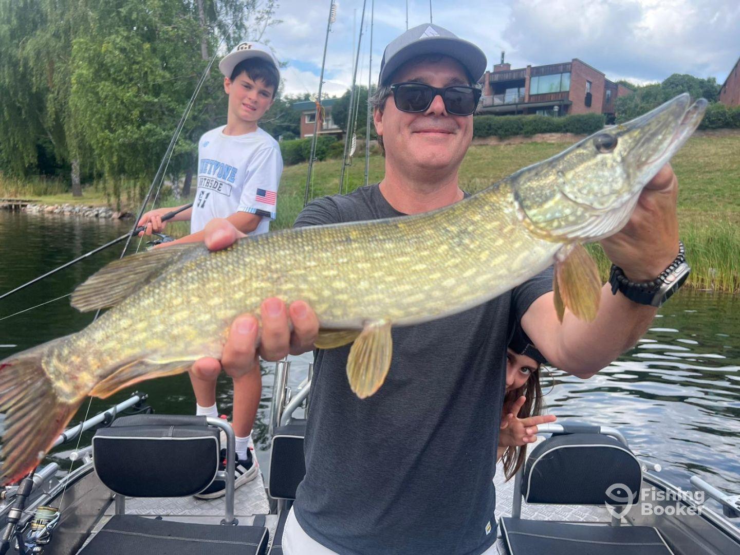 An adult angler proudly displaying a Northern Pike while two kids enjoy the fishing experience on a scenic lake.