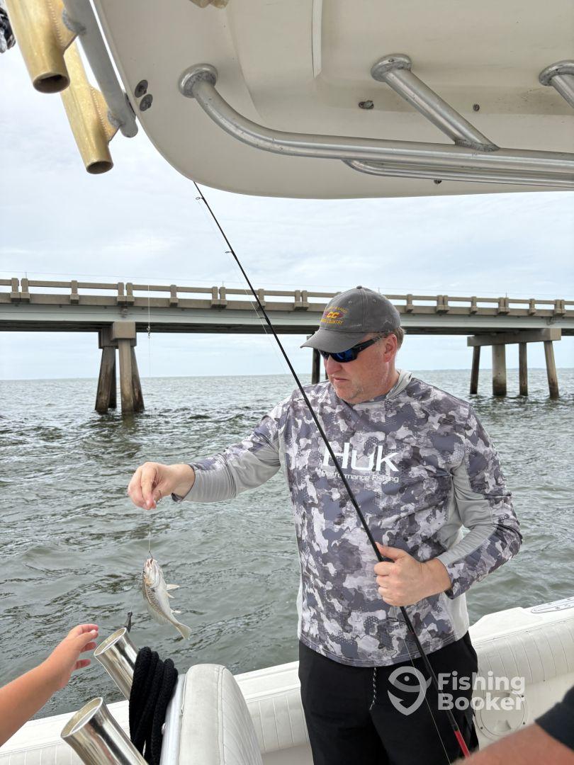 An angler reeling in a small fish while a child reaches out to assist, with a fishing pier visible in the background.