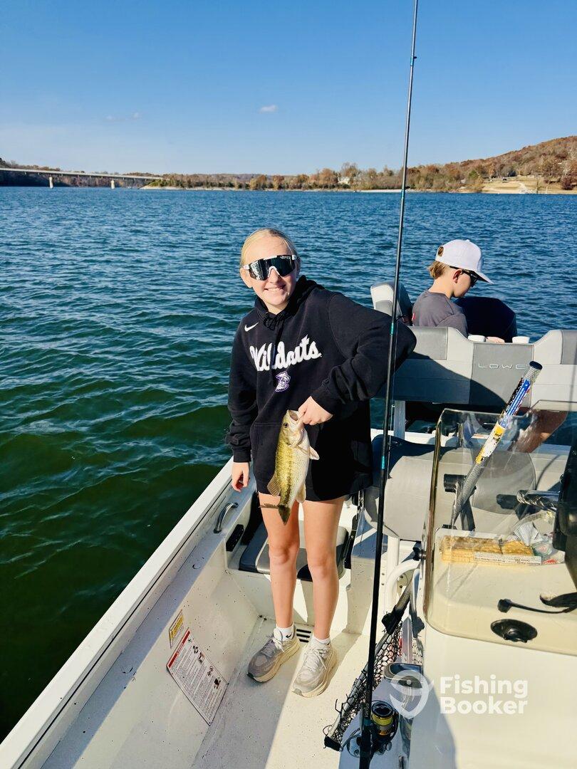 Young angler proudly holding a Largemouth Bass while fishing on a scenic lake, with a fellow angler in the background.