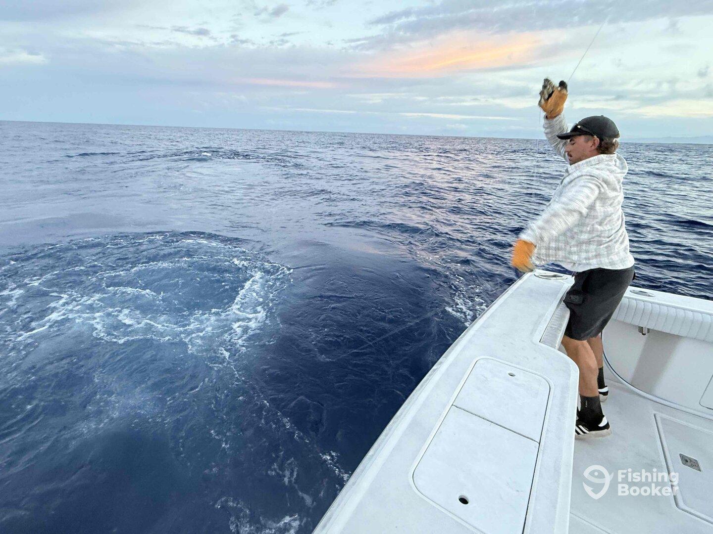 An angler casting a line from the bow of a sport fishing boat, surrounded by open ocean waters during a fishing trip.