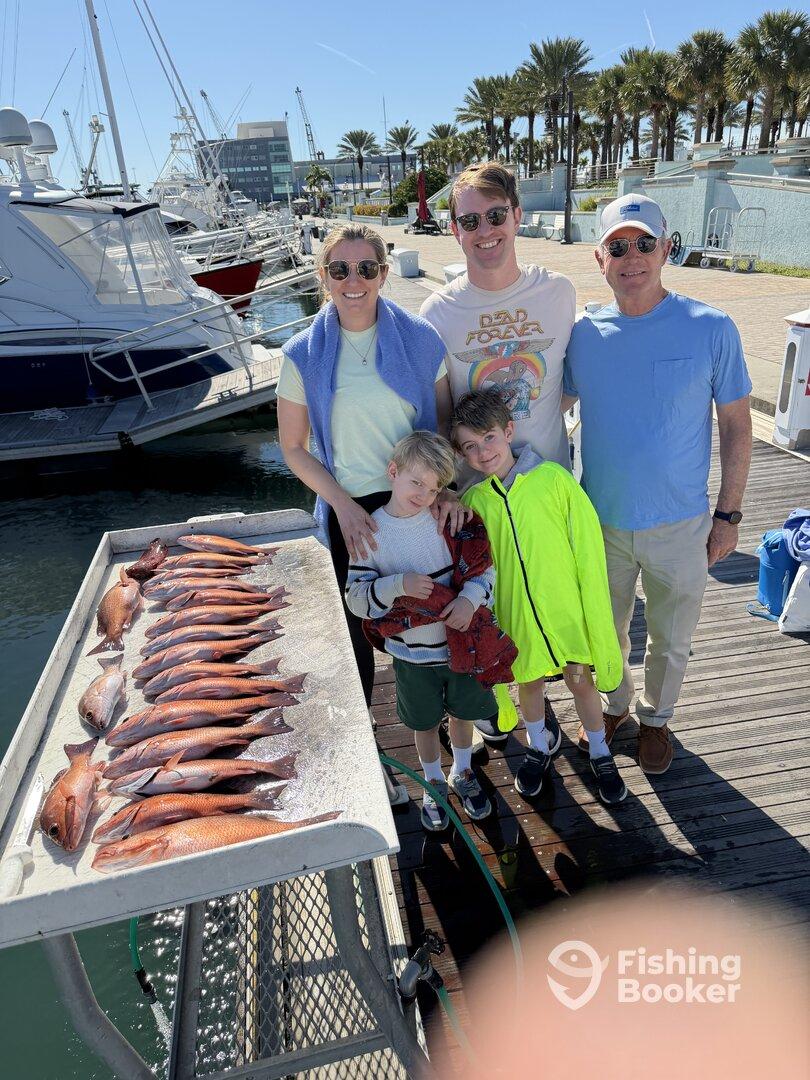 A family proudly displays their catch of Red Snapper on a dock after a successful fishing trip.