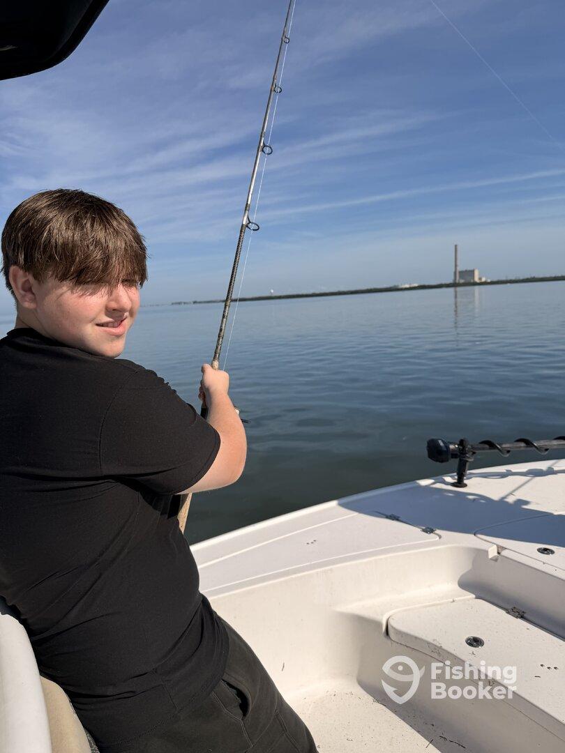 A young angler is reeling in a catch while fishing on a calm day, with a scenic backdrop of water and distant structures.