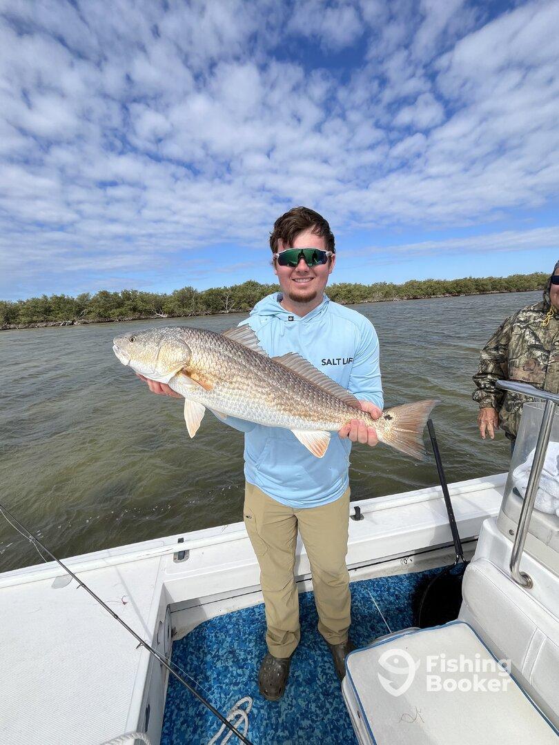 Angler proudly displaying a large Redfish while fishing from a boat in a scenic coastal environment.