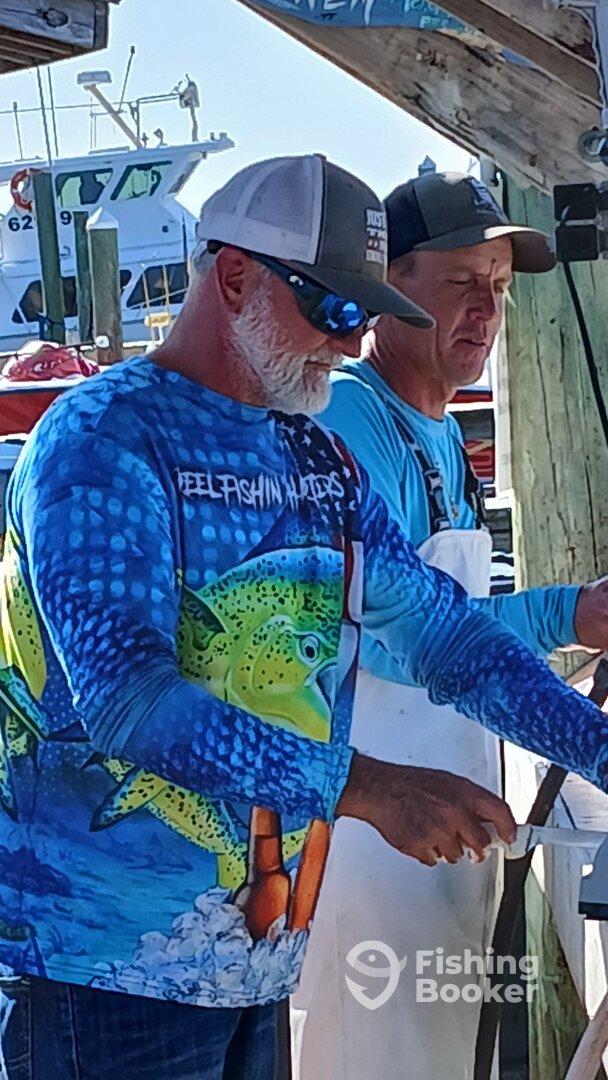 Two individuals engaged in a fishing-related activity at a dock, with boats visible in the background.