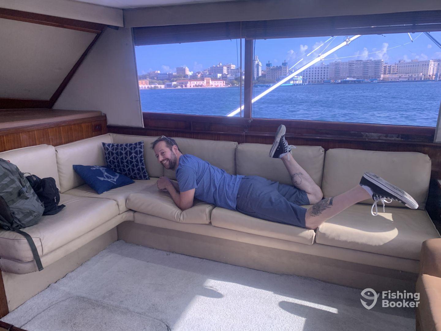 A relaxed angler lounging on the plush seating inside a spacious boat cabin, with a scenic waterfront view in the background.