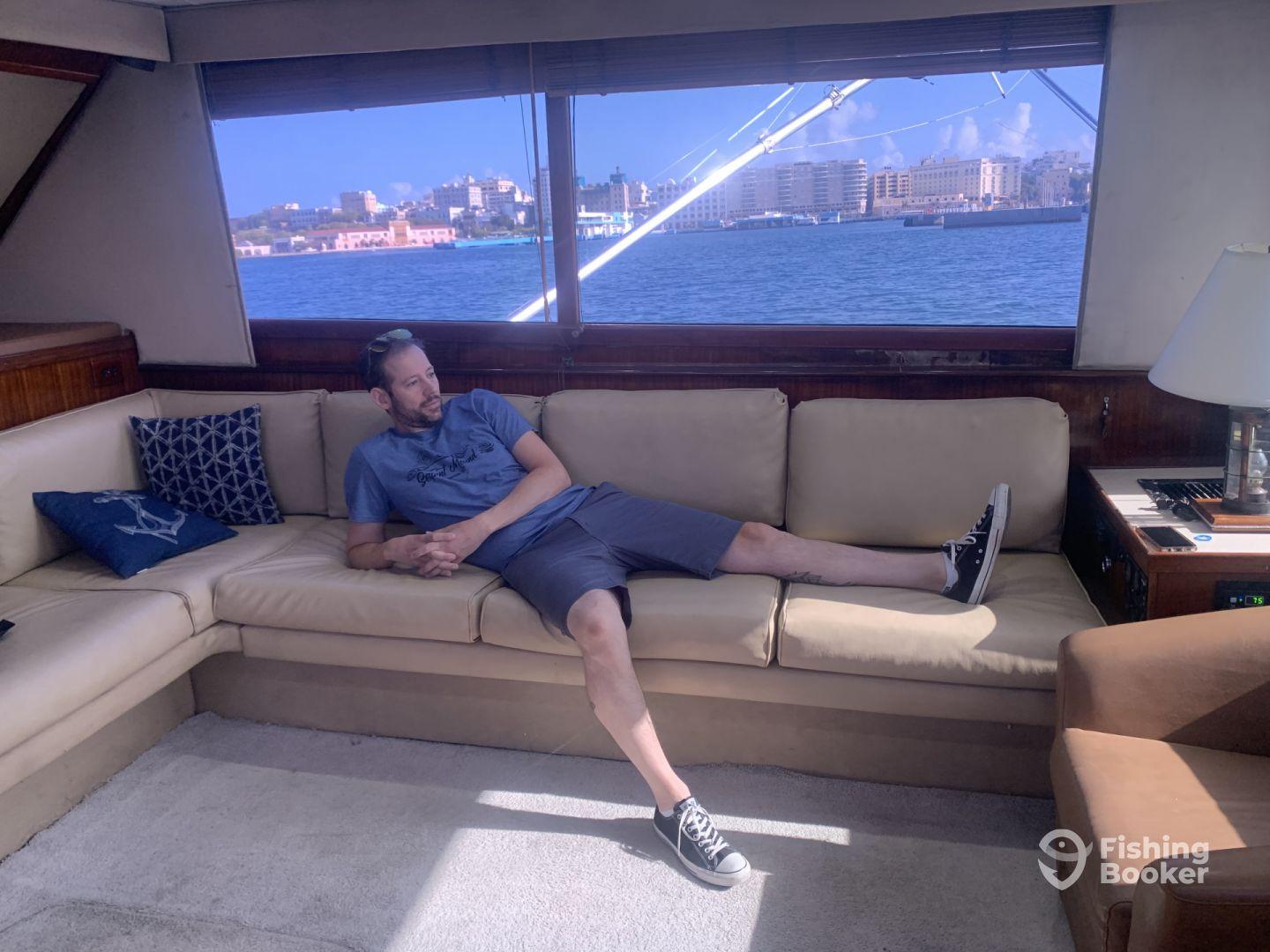 A relaxed angler lounging on the plush seating of a yacht's cabin, with a scenic waterfront view in the background.