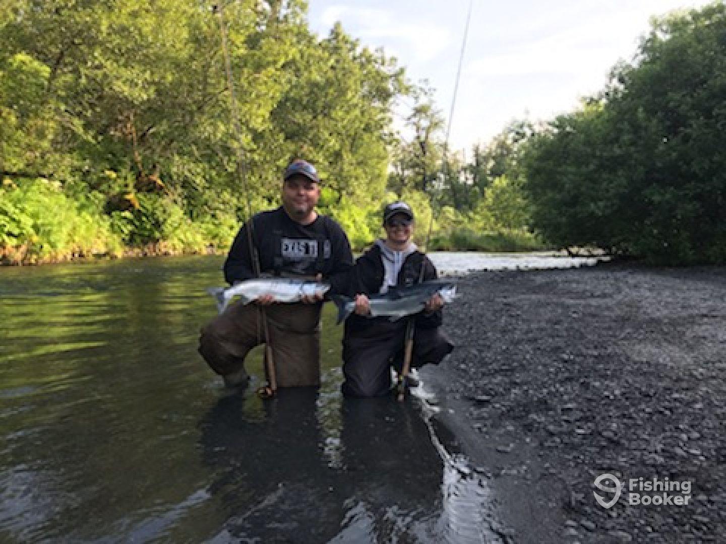 Two anglers proudly displaying their catches of salmon while wading in a scenic river, surrounded by lush greenery.