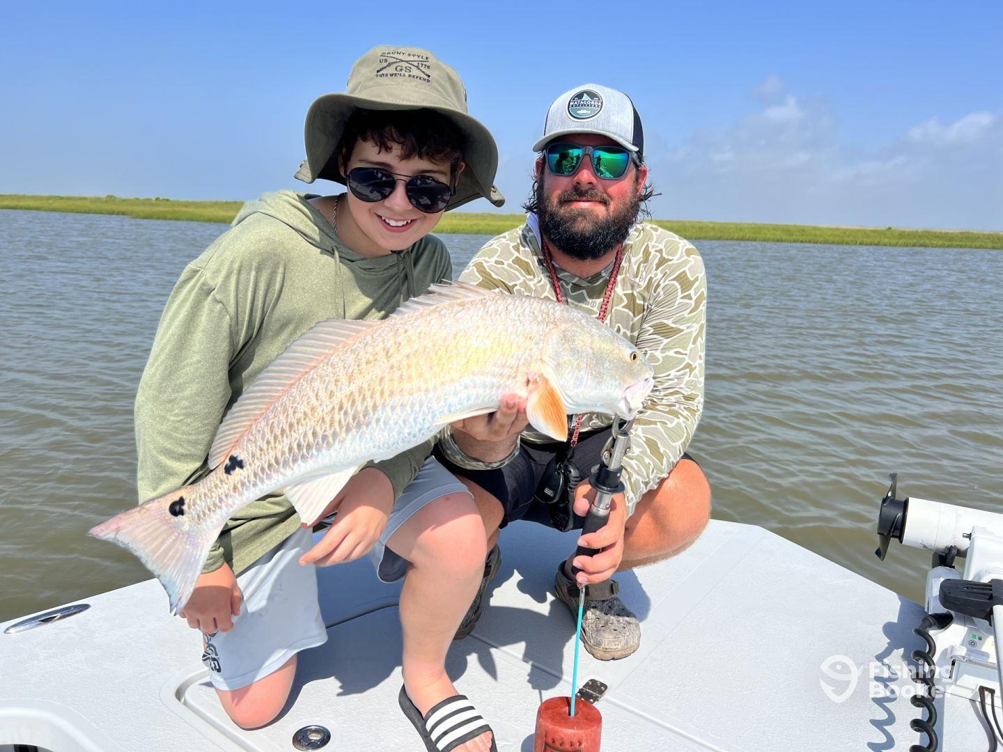 A young angler and an adult proudly displaying a large Redfish while fishing in a scenic coastal environment.
