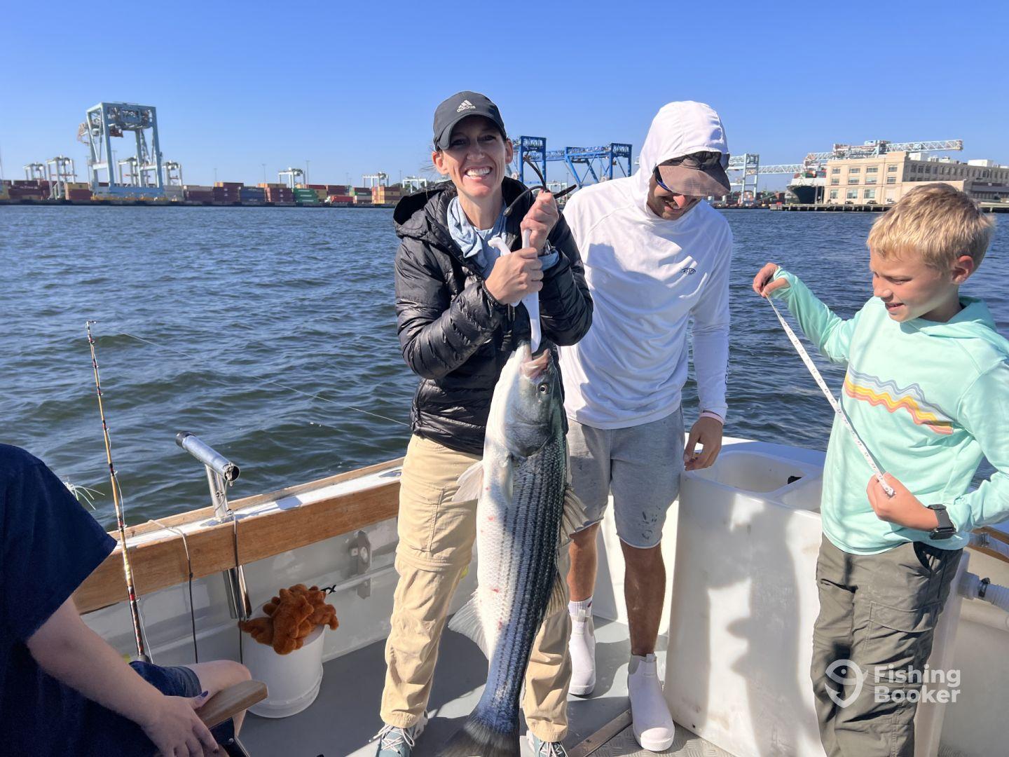 A group of anglers celebrating a successful catch of a large striped bass while fishing on a boat, with a bustling harbor in the background.