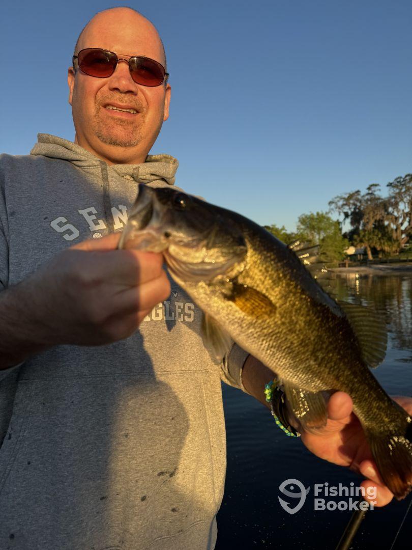 An angler proudly displaying a large Largemouth Bass while fishing at a serene lake during sunset.