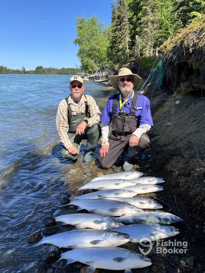 Two anglers posing beside a neat display of freshly caught salmon on the shore of a scenic river, showcasing their successful fishing trip.